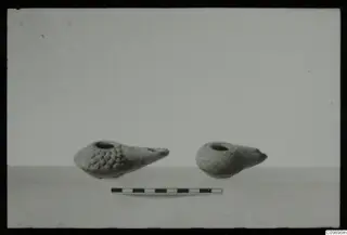 Two oil lamps with a measuring scale bar in front of them on a pale surface.