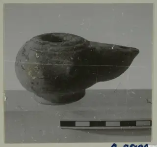 Oil lamp with a pointed nozzle and ring base, photographed in side view beside a scale bar.