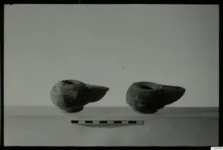 Two dark oil lamps with pointed nozzles stand beside a scale bar on a pale surface