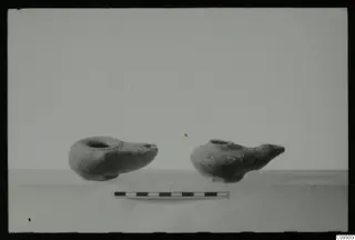 Two clay oil lamps with spouts, placed on a plain surface beside a scale bar
