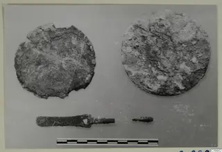 Two corroded round metal plaques and a metal tool on a grey backdrop, with a scale bar below; finds from Ajii Omologitades and Politiko