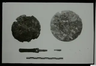 Two round, corroded discs, a rod-like tool and a small rod fragment on a white card, with a scale bar below.