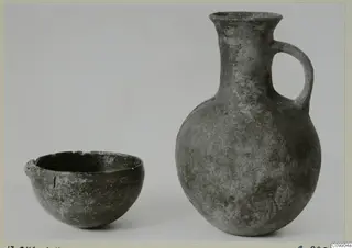 Dark grey pottery bowl and jug with a loop handle, standing side by side against a plain backdrop.