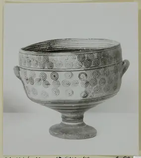 Footed bowl with two small handles and rows of concentric circles on its body, photographed against a plain backdrop