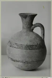 Pottery jug with a flaring neck, single strap handle and dark painted horizontal bands, photographed against a grey backdrop.