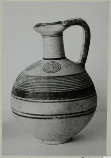 White Painted IV jug with a round body, tall neck and strap handle, decorated with dark bands and concentric circles