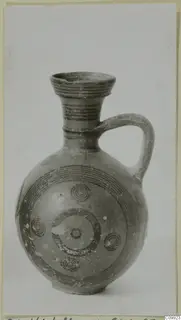 Round-bodied jug with a flaring mouth, a single strap handle and dark concentric circle painting, shown against a grey backdrop.