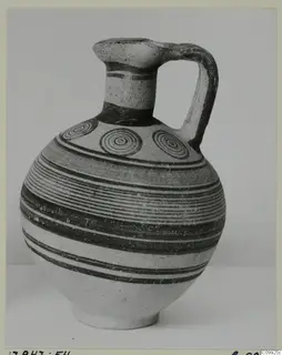 Jug with a strap handle, horizontal painted bands and concentric circles on the shoulder, photographed against a plain backdrop.