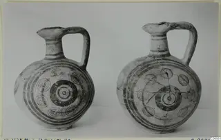 Two painted pottery jugs with concentric-circle decoration and single handles, photographed side by side against a grey backdrop.
