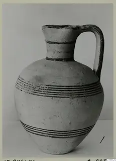 Pale pottery jug with a single strap handle and dark painted bands, photographed against a grey backdrop