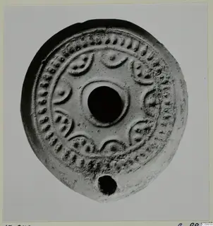 Round pale clay lamp seen from above, with a central filling hole and relief dots and crescent motifs around the rim