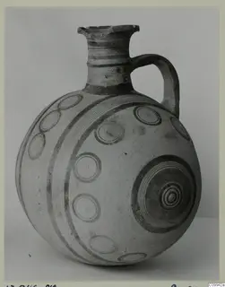 Round-bodied jug with one loop handle and dark painted concentric circles and bands on a pale surface.