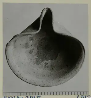 Terracotta lamp with a shallow rounded body and a pinched-up nozzle, shown against a grey background beside a scale bar.