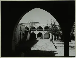 Whitewashed monastery courtyard with an arcaded building, a stone staircase and bare trees, seen through a pointed arch