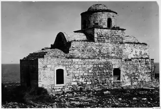 Monastery building of stone with a domed drum and arched windows, on rocky ground above the sea.