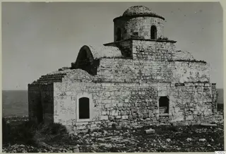 Monastery building of stone with a dome on a drum and arched windows, with the sea behind.