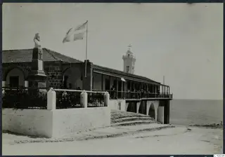Monastery building on the seafront with a flag, a cross and a bust on a pedestal