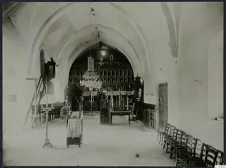 Vaulted monastery interior with a carved screen, a chandelier, a row of wooden chairs and a wooden door