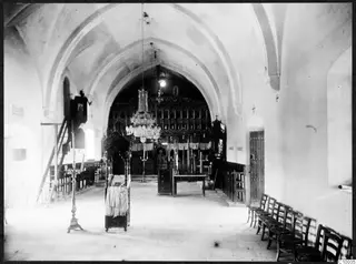 Interior of a monastery church looking toward a dark wooden screen with icons, with chairs along the right wall and a wooden door.