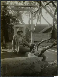 A smiling woman in a cloche hat sits on a large stone trough beside a dog, under a timber frame.