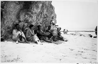 Group of people sitting against a rock face on a sandy beach by the sea.