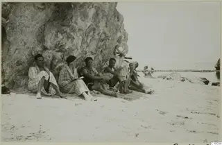 Group of seven people sitting against a rock face on a sandy beach by the sea.