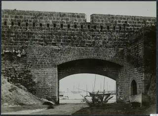 Arched gateway through a masonry fortification wall, with sailing boats moored in the harbour visible through the arch.