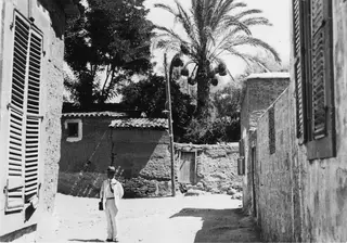 Ernst Kjellberg standing in a sunlit village street between mud-walled houses, with a palm tree behind.