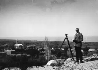 John Lindros beside a tripod-mounted instrument on a rocky hilltop above a village and plain