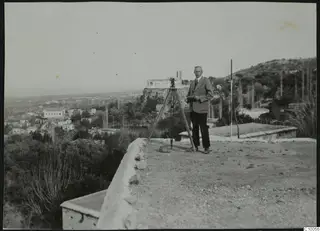 John Lindros standing beside a surveying instrument on a tripod, on a terrace above Lapithos