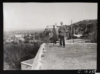 John Lindros standing beside a surveying instrument on a tripod on a terrace above Lapithos
