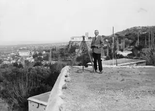 John Lindros standing beside a surveying instrument on a tripod on a terrace above Lapithos