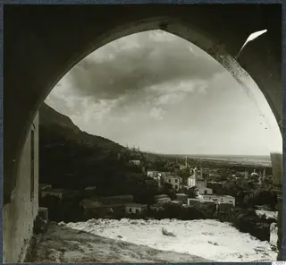 View through a stone arch over a hillside village of pale houses and trees, with a steep mountain slope on the left.