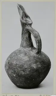 Dark grey-brown juglet with a tall trefoil-shaped spout and a single loop handle, photographed on a plain background.
