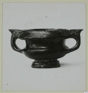 Black glazed pottery bowl with two loop handles and a short foot, seen from the side