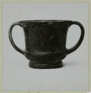 Two-handled dark glazed vessel with incised horizontal lines and a zigzag band on its body, seen against a plain background.