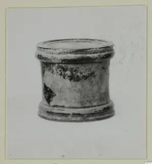 Small cylindrical vessel with a ridged rim and base and a dark, flaking glaze, shown against a plain background