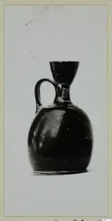 Small black-glazed jug with a single loop handle and a funnel-shaped mouth, photographed against a pale background
