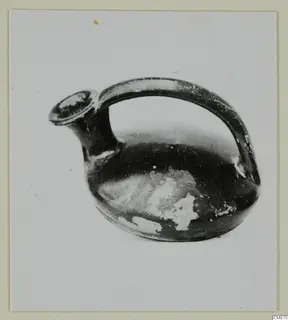 Black-glazed pottery jug with a spout and a strap handle, photographed on a plain background