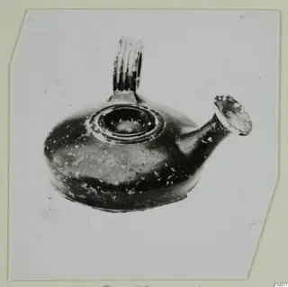 Black-glazed pottery oil lamp with a ribbed handle and a spout, photographed on a card mount