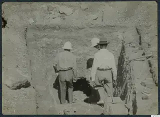 Three men in pale helmets and a dark hat walk toward a tomb entrance cut into excavated earth at Stylli.