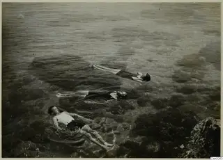 Three people floating face-up on clear, shallow sea water beside a rocky outcrop.