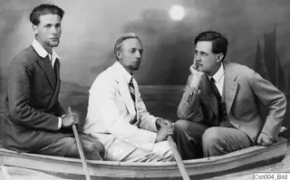 Three men posing seated in a rowing boat in front of a painted backdrop of sea, a moon and sailing boats.