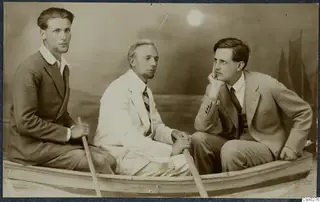Three young men posing seated in a small rowing boat against a painted studio backdrop of sea and sails.