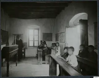 Children seated on wooden benches in a school room, with a bearded man in a long dark robe standing at the left.