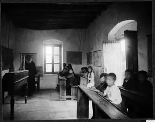 Classroom with a bearded teacher in a long dark robe standing by a desk, and children seated on wooden benches.