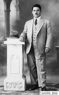 Studio portrait of Toulis in a light suit, standing beside a white pedestal with a hat on top