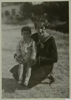 Woman kneeling on dry ground at Lapithos, holding a small boy on her lap, both facing the camera.