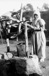 Woman in a light headscarf and long robe standing by a stone well with a wooden windlass and bucket