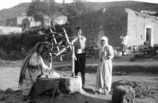 Three villagers beside a stone well with a wooden frame, in a village street with stone houses behind.